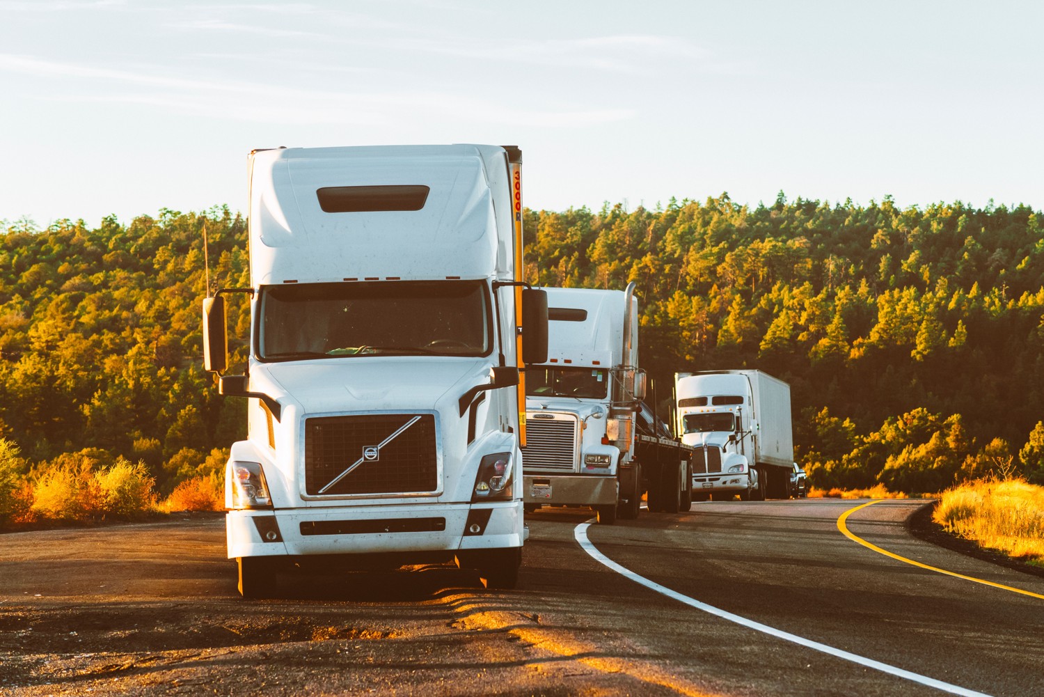 White conventional freight trucks on a road beside wooded hills